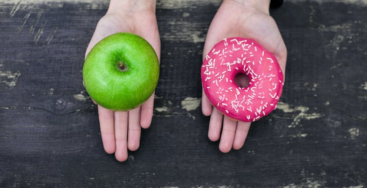 Two hands, one holding a green apple and the other holding a donut with pink icing and sprinkles