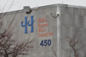A building with corrugated steel siding is seen on a snowy, grey day. On the side of the building is a blue H with a wave through it, Halifax Water's logo. Next to the H, written in orange, "Halifax Regional Water Commission." Under that, the number 450.