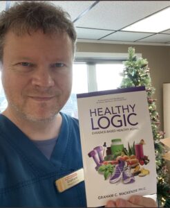 A man with wavy hair wearing blue scrubs and holding a book called Healthy Logic.